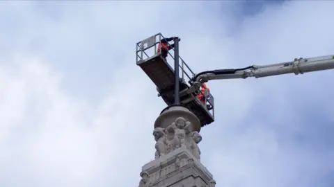 Engineers in hi-vis vests and hard hats work on an aerial platform on top of a clock tower to install the time ball. A carving with cherubs on it holds the plinth.