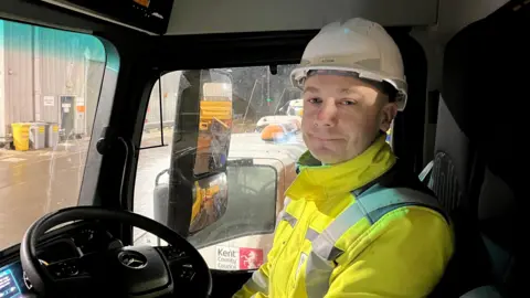 Phil Harrison/BBC Un hombre con chaleco amarillo de alta visibilidad sentado en la cabina de un camión con un casco blanco.