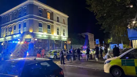 Police officers stand behind a cordon on a residential street in Clapham at night, with forensic tents and emergency vehicles visible near a pub as investigators examine the scene of a fatal shooting.