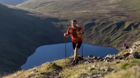Nick Moore Nick Moore on a mountain ridge. He is wearing a red top and black shorts and carrying a yellow rucksack and walking poles. Behind him is a tarn, reflecting the blue of the sky and ringed by further hills. The head and shoulders of his companion, who is wearing a pink peaked cap, can be seen ascending the ridge behind him.
