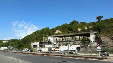 The derelict site, which has the remainder of a building, there are cars in front of it from across the road, you can see the clifface in the background.