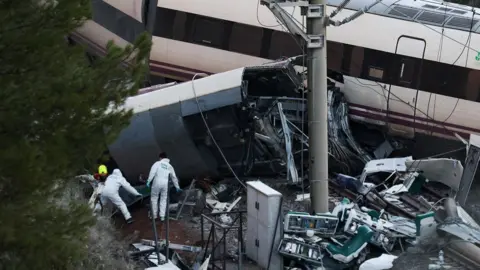 the scene of a train crash. A derailed carriage can be seen as police officers walk on the rails and embankment around it.