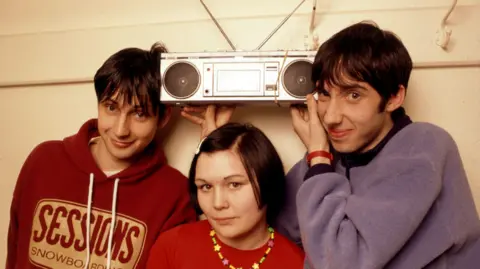 Getty Images The band Bis - John Clark, Amanda McKinnon and Steven Clark in 1995, holding up a record player 