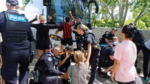 EPA/Shutterstock Protesters stand and sit in the street in front of a bus, with police present 