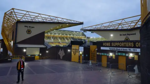 The orange and black Molineux is photographed in the dusk with lighting on and the entrance for home supporters in view. A fan is seen walking away from the ground with a scarf on his shoulders.

