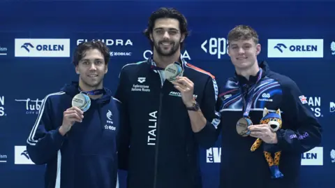 Wojtek Jargilo/EPA/Shutterstock Three men, all wearing dark tracksuits from their national swimming teams (France, Italy and GB) stand on a medal podium holding up their European Aquatics medals. Oliver Morgan on the right is holding a soft toy mascot with black spots on yellow skin, and wearing a blue bandana, shorts and boots. 