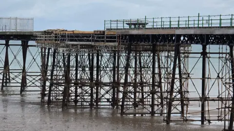 A close-up of a section of the iron pier, with the central section surrounded by scaffolding. There is fencing on the platform. At the base, the large rods meet a sandy beach and a calm sea on a cloudy day.