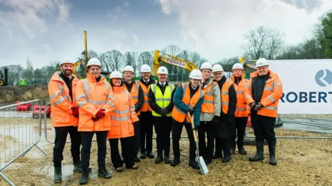 Bradford Council A group of people on a building site. They are wearing fluorescent jackets and wearing hard hats. They are smiling for the camera. 