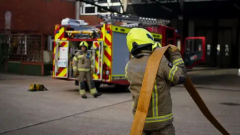 PA Media A stock image showing two firefighters in full uniform. One stands by a fire engine looking towards the camera and is slightly blurred. The other looks away from the camera and is in the foreground holding a fire hose