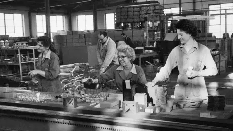 Historic England Black and white photograph of three women working at a table with lots of objects on it. The women are wearing jumpsuits and smiling. The middle woman is older and has grey curly hair and black glasses. In the background is a man picking up an object from a metal basket. 