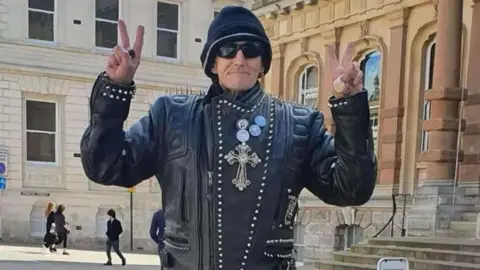 A man, William McNicholl, is pictured outside the Town Hall in Ipswich. He is wearing a black leather jacket and making 'peace' signs with both of his hands. He's also wearing sunglasses and a hat