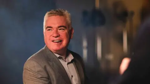 Daniel Lewis/Zurich Lee Michael Walton, a man with short grey hair wearing a grey suit jacket and grey shirt, sits at a black grand piano looking off to the side of the camera and smiling