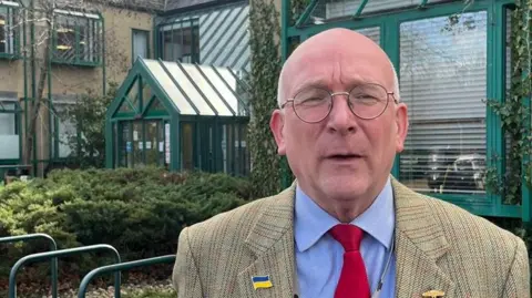 Richard Keeling is wearing a light blue shirt, a red tie and a beige blazer with a Ukraine flag on the lapel. He is wearing glasses and standing at the entrance to Teignbridge District council offices.