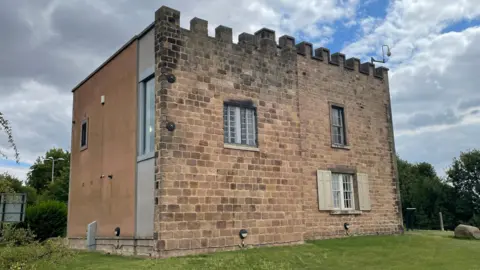 A stone-built structure with castle-like battlements stands on a grassy area beneath a cloudy sky. The building combines traditional masonry on the front with a more modern extension on the side, creating a blend of historic and contemporary architecture. Tall windows, shuttered openings, and surrounding trees give the scene a quiet, rural atmosphere.