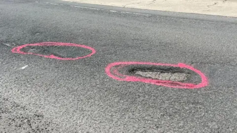 Two potholes on a road which have been marked up by pink spray paint circles.
