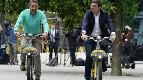 PA Media Chris Boardman in light green shirt and khaki trousers cycles alongside Andy Burnham who is dressed in a blue suit with a white open necked shirt