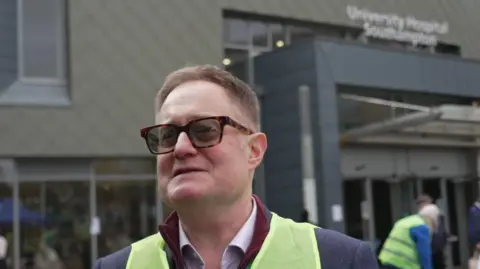 Head and shoulder photo of a man in hi-vis over a suit stands outside a hospital 