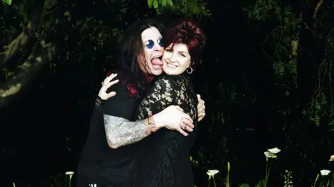 Steve Read Ozzy and Sharon Osbourne pose together outdoors in front of foliage, with their arms around each other. Ozzy Osbourne is wearing sunglasses and has tattoos on his arm and is pretending to lick Sharon's face. There are plants and flowers around them.