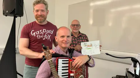 Jon Wright/BBC Two men stand next to each other, with a third sat in front, holding an accordion and a ukulele.