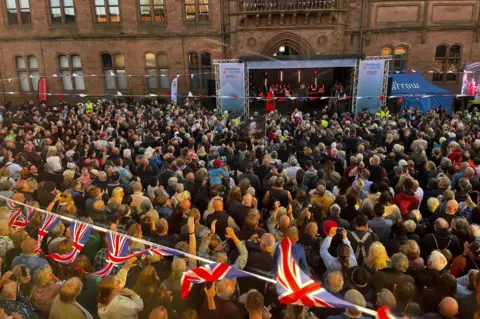 Crowds watching Si King and Lili Myers give a speech from an outdoor stage in Barrow. Union flag bunting is draped across the street.