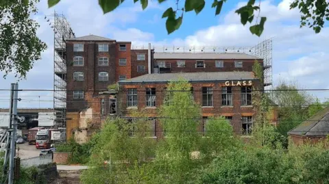 Stephen Hartland A brown brick building with scaffolding at the side. At the front of the building is a metal fence and some green trees. The word 'Glass' is at the top of building. 