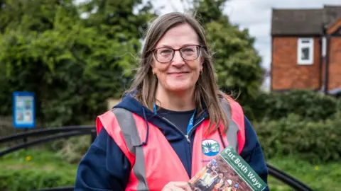 Lichfield and Hatherton Canals Restoration Trust A woman with long fair hair and dark-rimmed glasses smiles at the camera. She wears a pink high-visibility vest over a navy hooded top carrying the Lichfield and Hatherton Canals Restoration Trust logo. She holds a booklet titled 'Cut Both Ways' and stands in front of trees and a red-brick house.