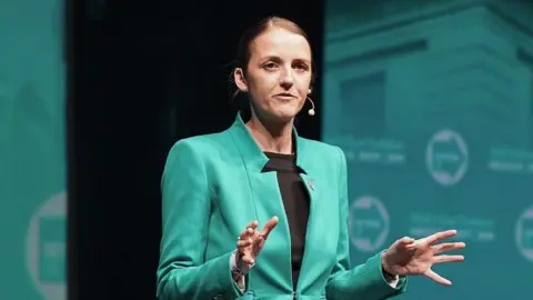 Getty Images Maria Bowtell dressed in a turquoise suit, matching the colour of the Reform UK backdrop, addresses a party conference. Her hair is scraped back and she is making a point with her hands.