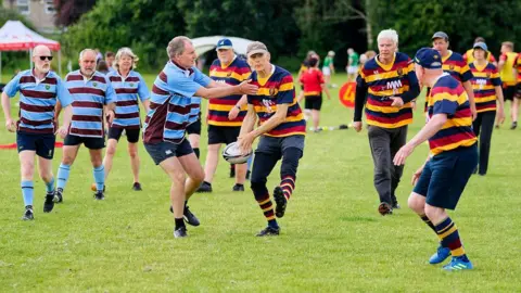 Eamonn Twamley Players take part in the Kingswood Walking Rugby Festival, with one team wearing blue and brown striped tops and one in a more colourful yellow, red and blue strip. A man is being tackled as he passes the ball