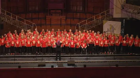 BBC/The One Show Over 200 members of the choir are dressed in red T-shirts on a large stage. The conductor is dressed in black and has bright purple hair and raised arms.