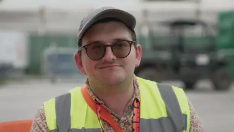 William Ogden looking to camera. He has on black rimmed glasses that are tinted and a cap. He is wearing a high-vis jacket.