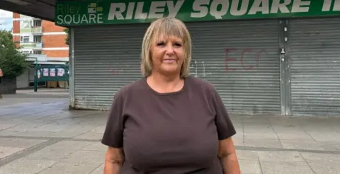 Julie Byrne, who has shoulder-length hair with blonde highlights is standing in Riley Square, with a shuttered shop behind her, along with paving.