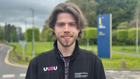 Chris Murray is standing in front of a blue sign that says Ulster University. He is wearing a black jacket with the initials UUSU on it in white and pink writing. He has dark wavy hair and a dark beard and is smiling into the camera.