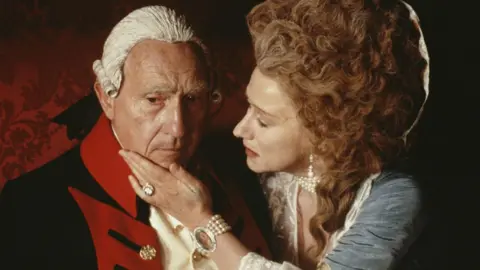 A film still showing two actors playing the roles of an 18th Century king and queen. On the left, a man wears a white wig and black and red military jacket over a white shirt. He has a pensive look on his face. On the right, a woman wearing a ginger bouffant wig, blue dress and pearls who holds the man's chin in her hand.