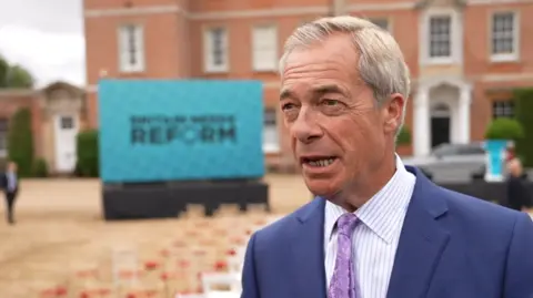 Nigel Farage looks towards a reporter off camera, with his mouth open as he talks, while wearing a blue suit with a striped shirt and purple tie, with chairs outside a stately home blurred in the background