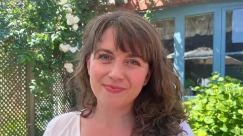 Daisy, who wears a white top and has shoulder length brown hair. She is photographed outside in her garden