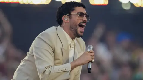 EPA Bad Bunny in dark glasses, a beige suit, and cream shirt and tie singing into a microphone on stage during a concert in Barcelona on 22 May 2026