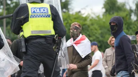 PA Media A police officer stands in front of two masked men