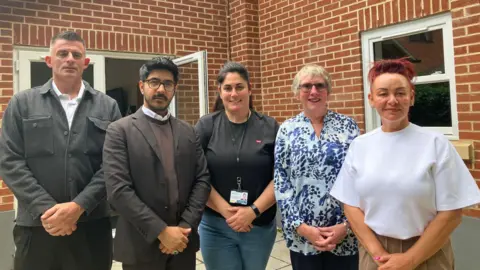 JOANNA TAYLOR/BBC Michael Wright and Abdul Khan from My Housing CIC alongside Lorna Thorley, Alison Jones and Kelly Slack from Peterborough City Council stand in a line, smiling at the camera. Behind them is a red brick wall with windows.