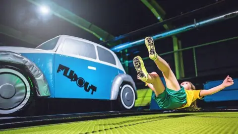 Image showing green landing mat and blue foam car with "Flip Out" written on the side. A child in a yellow T-shirt and green shorts is landing on the mat.