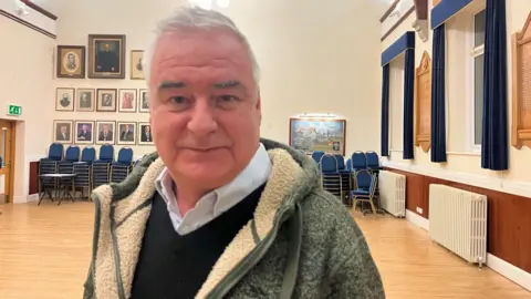 A man in a grey jacket smiling at a camera in a town hall.