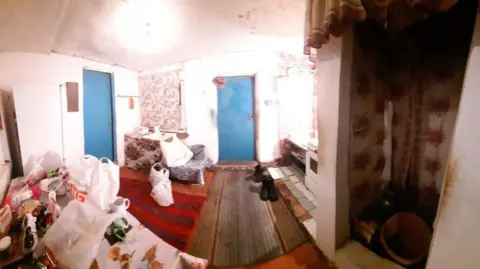Viktoriia Izviekova Image shows the accommodation Viktoriia and her mum stayed in whilst in Poltava. In the photo is one big room. There are two doors which are sky blue in colour. The floor on one side of the room is wooden and covered in a rug. The rug is red in colour. The other side of the room is tiled on the floor. The tiles are brown, green and white. There is a small separate area in the right hand side of the picture which has an orange bucket on the floor. The walls in the rest of the room are white. There is a small chair and a table on the far side of the room. On the side closest to where the photo was taken, there is a long table. It is covered in carrier bags with shopping. there is a mug on the table too. 