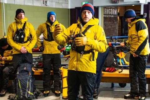 Angus Trinder/Cairngorm Mountain Five CMRT volunteers - one of the group is partly hidden by the volunteer nearest the camera - stand inside a building at Cairngorm Mountain. The volunteers are wearing yellow and black jackets, black trousers and woollen hats.