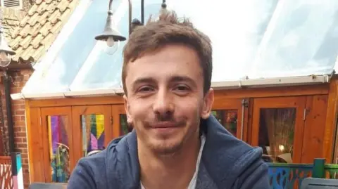 A smiling man sat down in an outdoor location. He has brown hair and a short beard and is wearing a blue hooded top. 