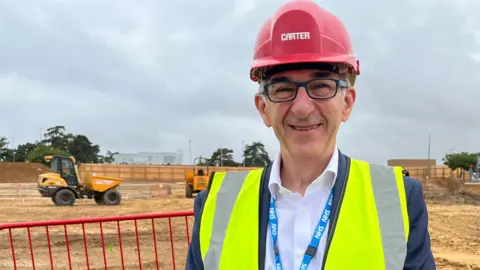Clare Worden/BBC Peter Cox stands next to the construction site of the new car park. The bare sandy soil is exposed. He wears a red hard hat and a yellow high-vis waistcoat.