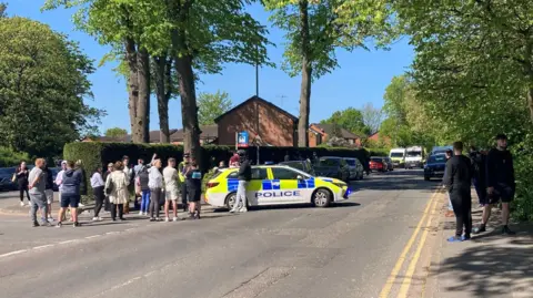 A police car parked in the middle of a residential road with a group of people standing by the car speaking to a police officer.