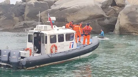 Taiwanese coast guard personnel dressed in orange uniforms on board a coast guard ship apprehend a suspect dressed in a blue top and grey trousers, who appears to be kneeling on a small floating platform. The vessels are by large rocks.