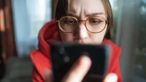 Woman wearing glasses looks at phone which is prominent in the foreground.