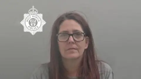 West Mercia Police A woman's mug shot. She has purple-red hair and is wearing large circular glasses and a long sleeve grey jumper on.