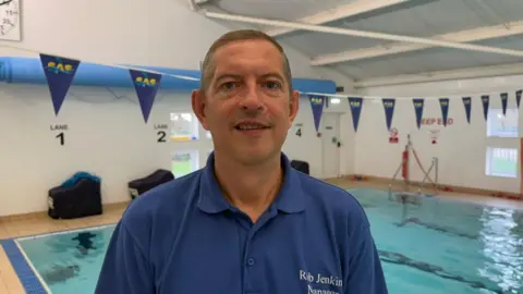 A man is looking at the camera. He's pulling a neutral expression. He's wearing a blue polo shirt. Behind him is a swimming pool, which has bunting hung above it.