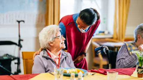 A woman with dark hair wearing a red tabard over a blue top is leaning over an elderly woman who is eating a plate of food at a table.
